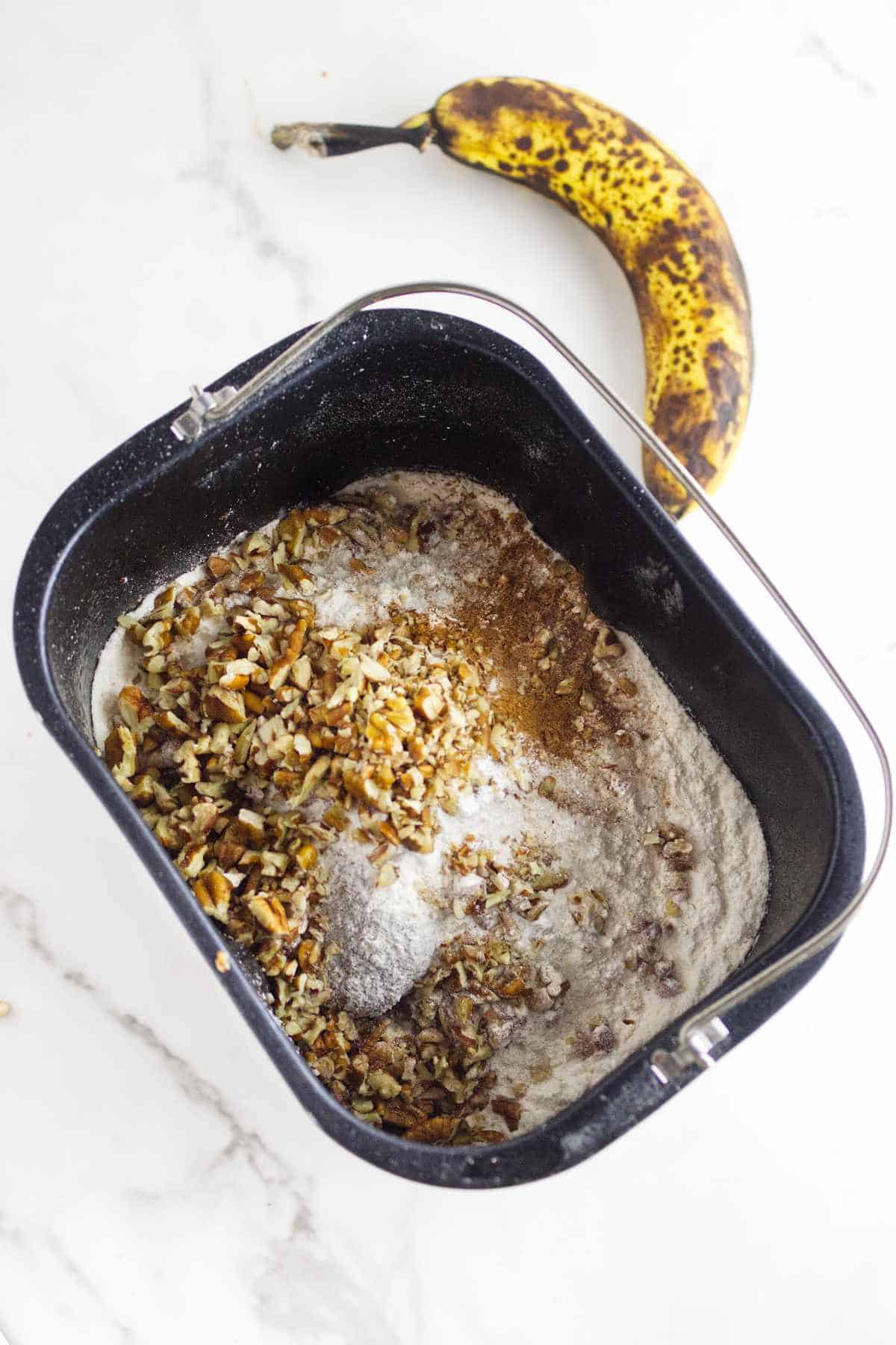A bread machine pan filled with flour, chopped nuts, cinnamon, and other ingredients for bread machine banana bread sits on a white surface next to a ripe, spotted banana.