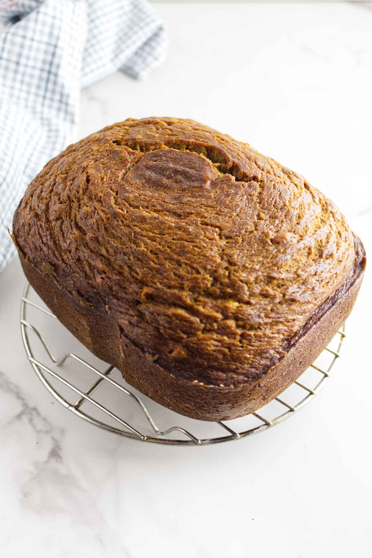 A golden-brown loaf of bread machine banana bread cools on a round metal rack atop a white marble surface, with a blue and white checkered cloth visible in the background.