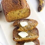 A loaf of bread machine banana bread sits on a cutting board with three slices cut, two topped with melting pats of butter. A ripe banana rests in the background.