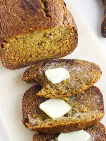 A loaf of bread machine banana bread sits on a cutting board with three slices cut, two topped with melting pats of butter. A ripe banana rests in the background.