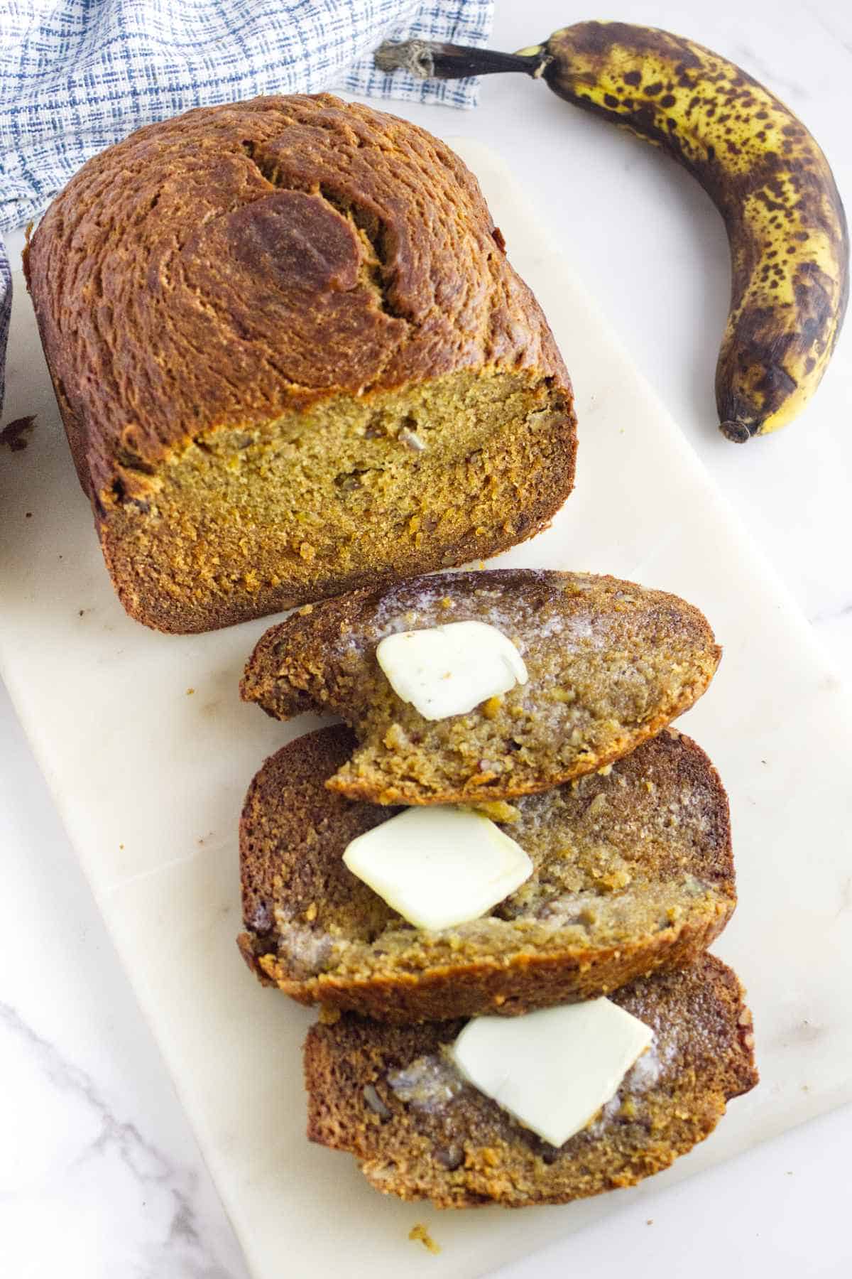 A loaf of bread machine banana bread with three slices cut, each topped with melting butter, sits on a white surface next to a ripe, spotted banana and a blue-checked kitchen towel.