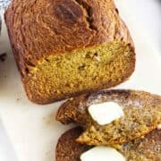 A loaf of bread machine banana bread rests on a white surface with two slices cut, one topped with melting butter. A ripe banana and a blue cloth are partially visible in the background.