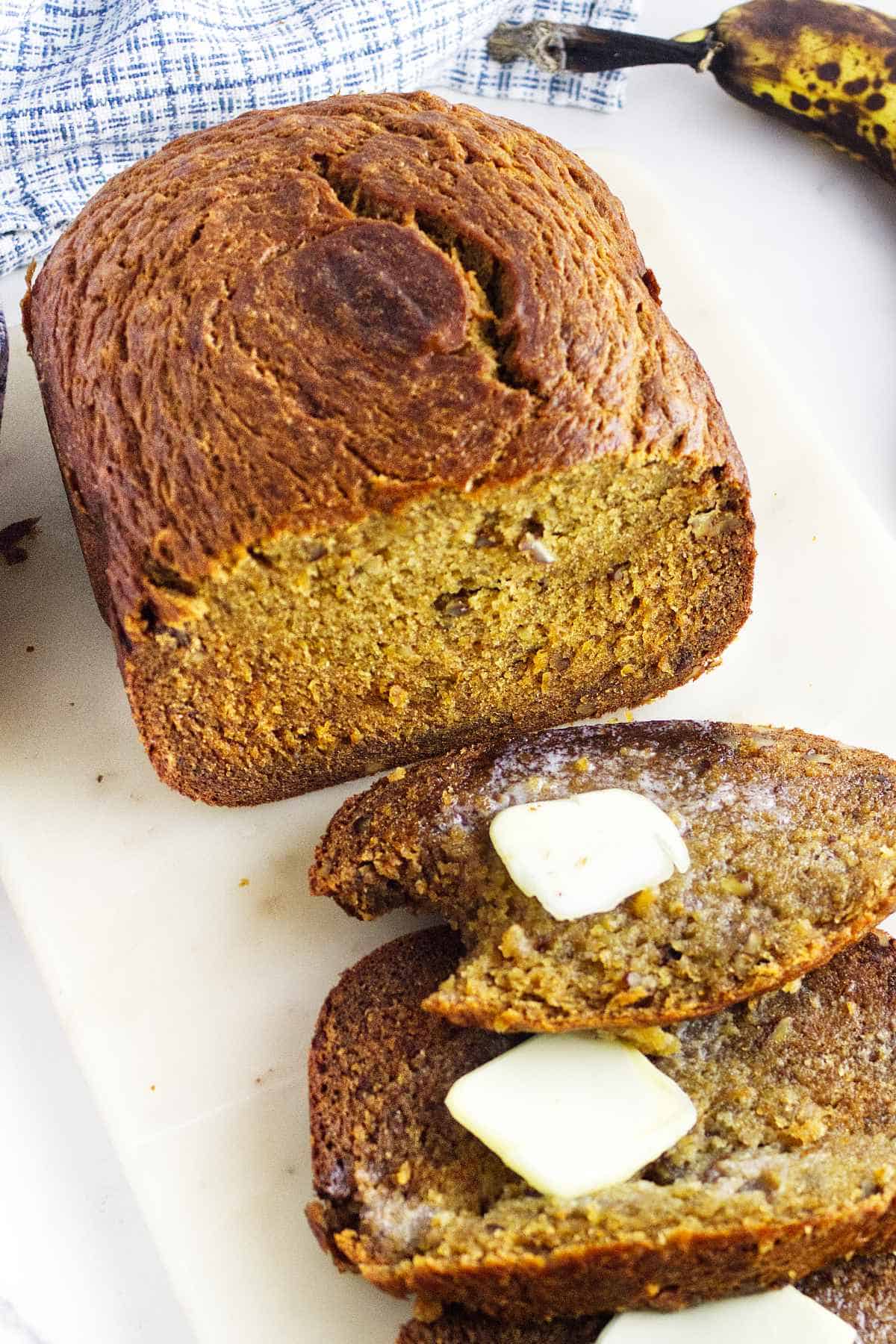 A loaf of bread machine banana bread rests on a white surface with two slices cut, one topped with melting butter. A ripe banana and a blue cloth are partially visible in the background.