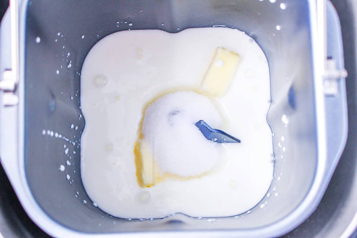 Close-up of a bread machine pan with milk, sugar, butter, and other wet ingredients for bread machine buttermilk bread before mixing. The paddle is visible in the center, viewed from above.