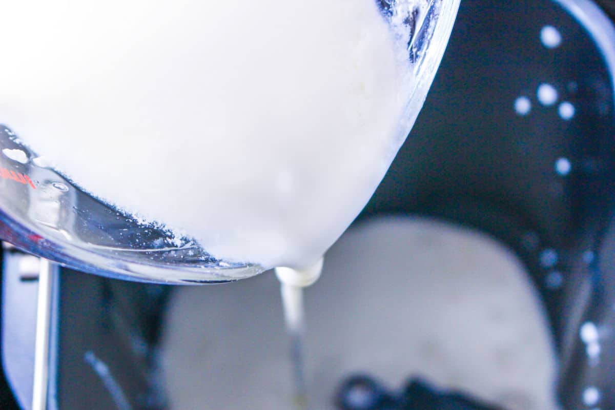 A close-up of milk being poured from a measuring cup into an ice cream maker, with the churning paddle visible inside the machine—perfect for pairing with homemade bread machine buttermilk bread.