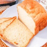 A loaf of freshly baked bread machine buttermilk bread sits on a white plate, with two slices cut and laid out in front. The bread has a golden-brown crust and a soft, airy interior.