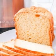 A loaf of homemade bread machine buttermilk bread sits on a white plate, with three slices cut and laid in front. The golden-brown crust and soft, airy interior are inviting, while a metal appliance and glass jars sit in the background.