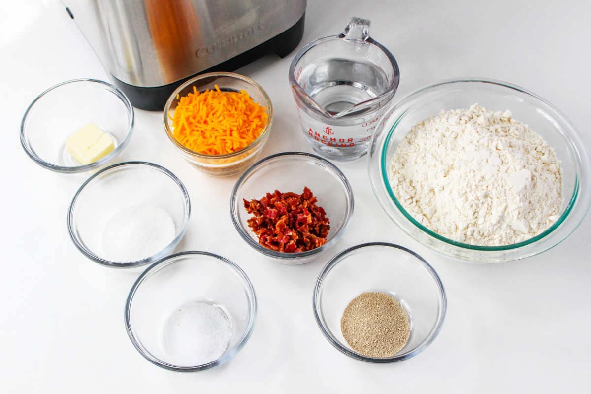 Eight glass bowls and a measuring cup hold ingredients for bread maker cheese bread-flour, shredded cheese, chopped bacon, yeast, sugar, salt, butter, and water-on a white countertop beside a stand mixer.