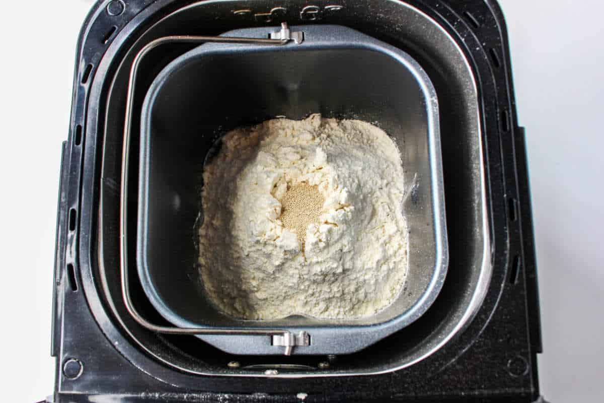 An overhead view of a bread maker pan filled with flour and a small mound of yeast in the center, ready to be mixed for delicious cheese bread.
