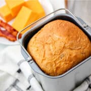 A freshly baked loaf of bread maker cheese bread sits in a pan on a kitchen towel, with slices of cheddar cheese and strips of bacon on a white plate in the background.
