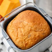 A freshly baked loaf of cheese bread rests in a bread maker pan on a checkered towel. In the background, several slices of cheddar cheese sit on a white plate.