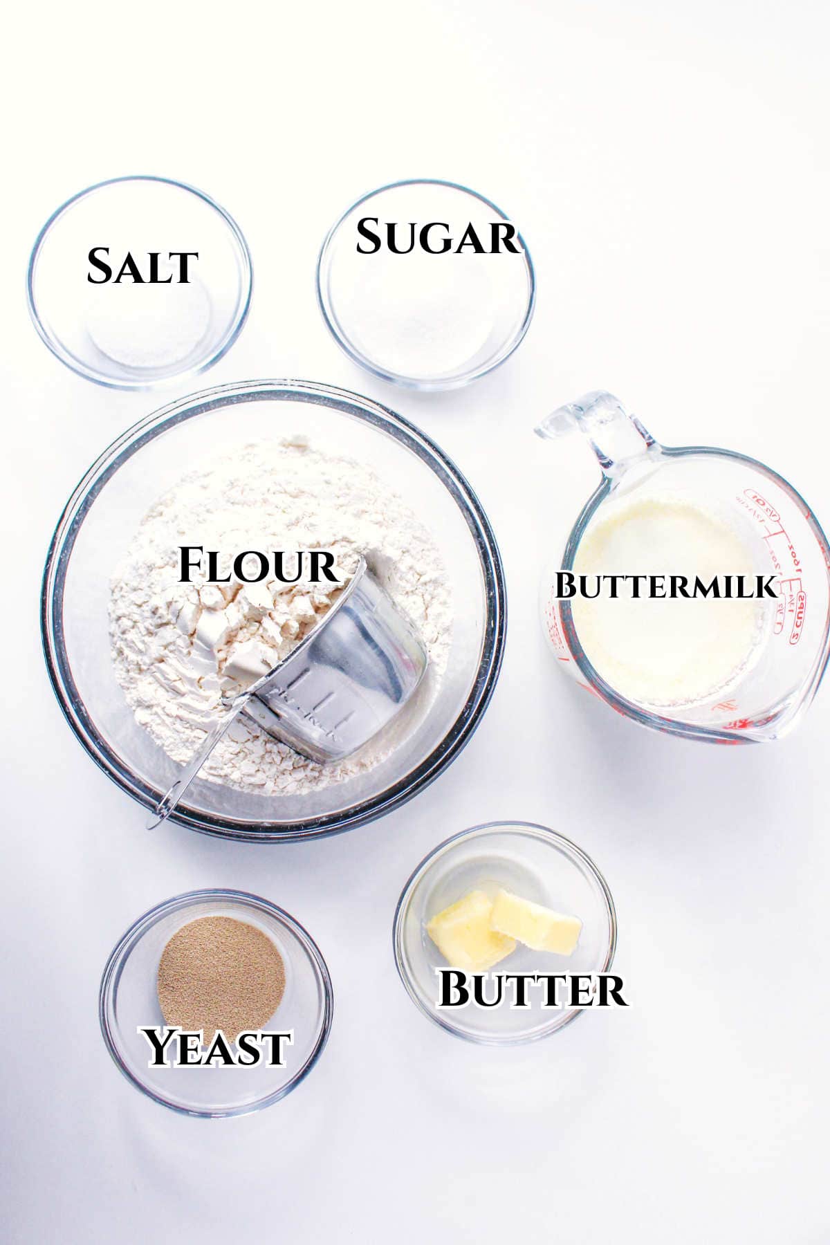 Six clear bowls showcase labeled baking ingredients for bread machine buttermilk bread: salt, sugar, flour (with a measuring cup), buttermilk (in a measuring jug), yeast, and two pieces of butter, all arranged on a white surface.
