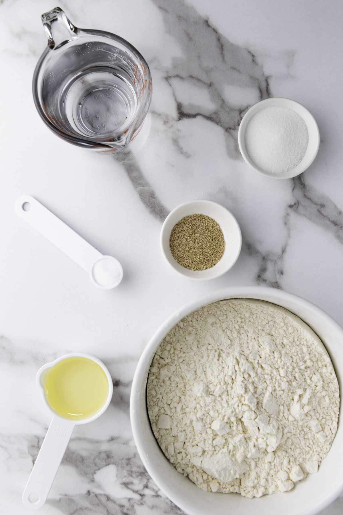 A marble countertop with bowls and measuring cups holding flour, water, sugar, oil, salt, yeast—everything arranged neatly to prepare bread machine honey wheat bread.
