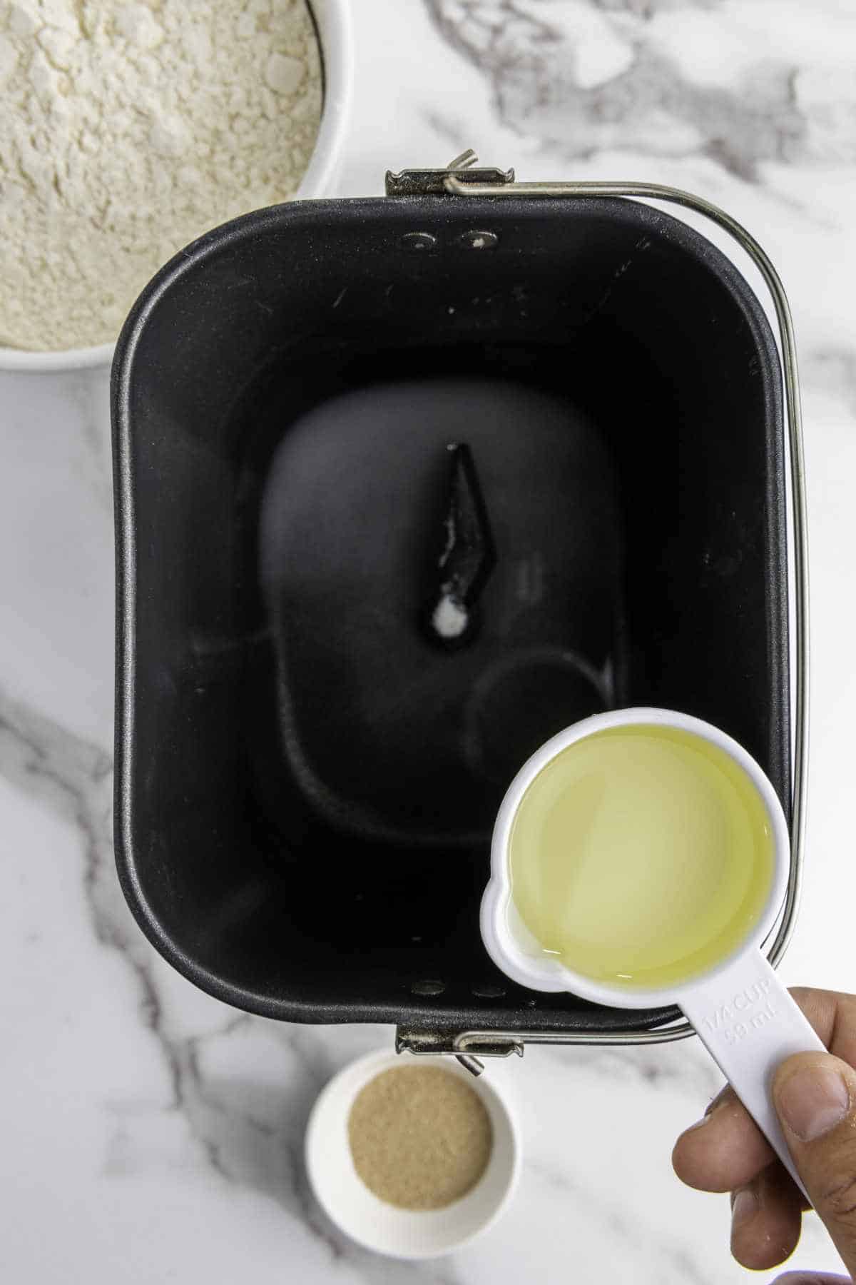 A hand holds a white measuring cup of oil over the empty pan of a bread machine, ready to start making honey wheat bread. Flour in a bowl and a small bowl of yeast sit on the marble countertop nearby.