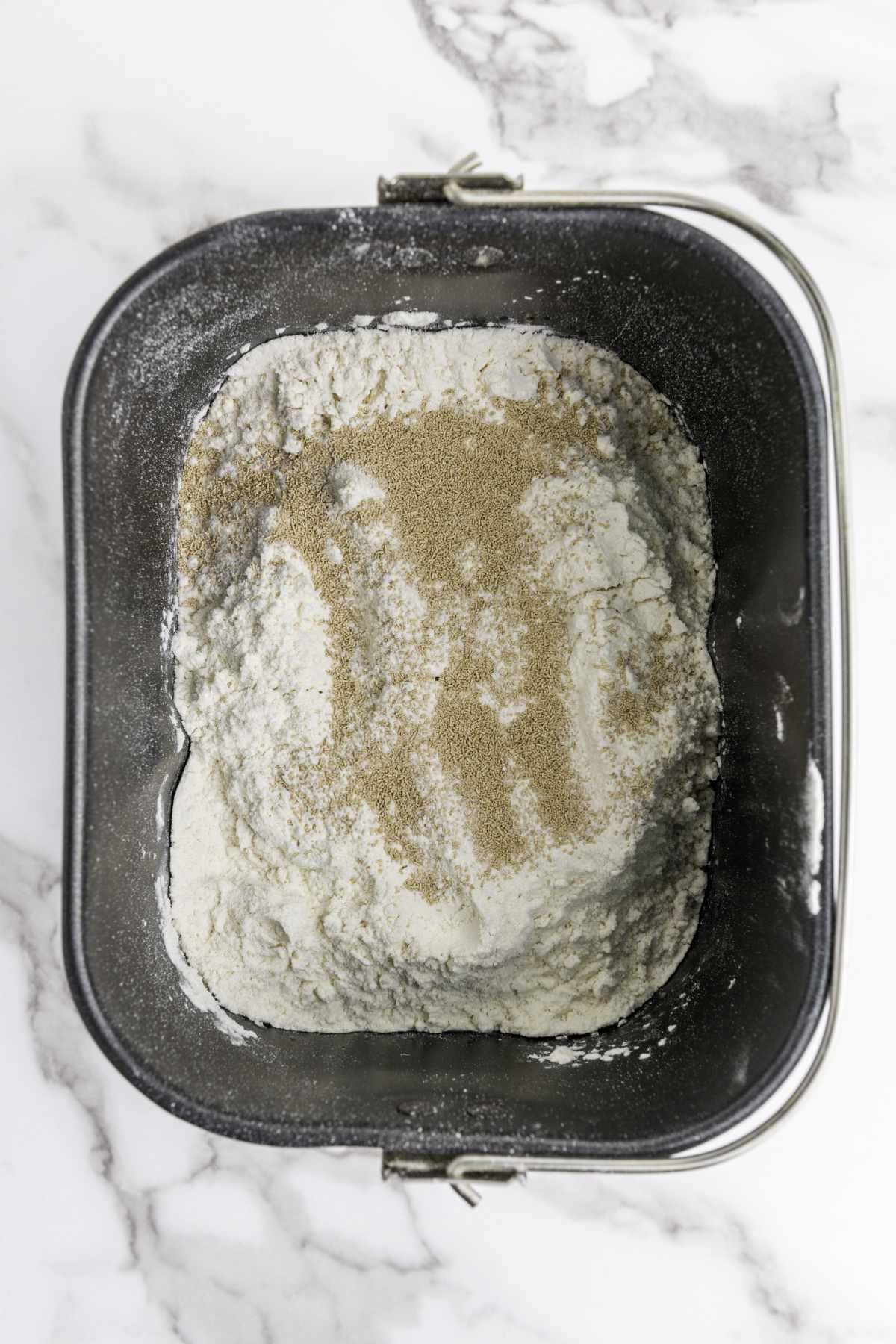 A bread machine pan filled with flour and a layer of dry yeast on top, ready for baking delicious honey wheat bread, sits on a white marble surface.