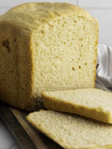 A loaf of bread machine honey wheat bread sits on a wooden board with two slices cut and lying in front. The bread has a golden-brown crust and soft, airy texture. A striped cloth is partially visible in the background.