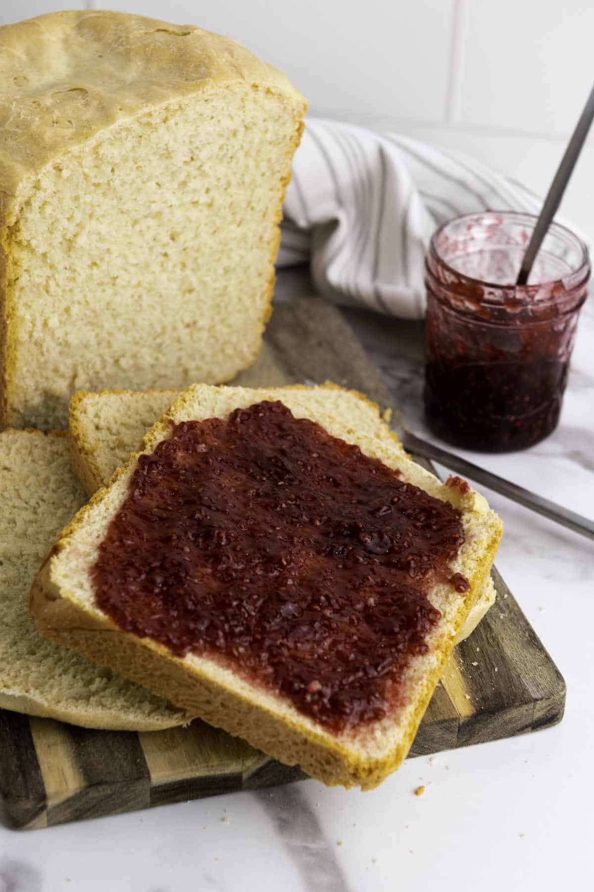 A slice of bread machine honey wheat bread spread with raspberry jam sits on a wooden cutting board beside a loaf and an open jar of jam with a spoon. A striped cloth is in the background.