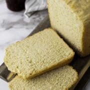 A loaf of bread machine honey wheat bread sits on a wooden board with two slices cut, next to a jar of jam and a striped cloth in the background.