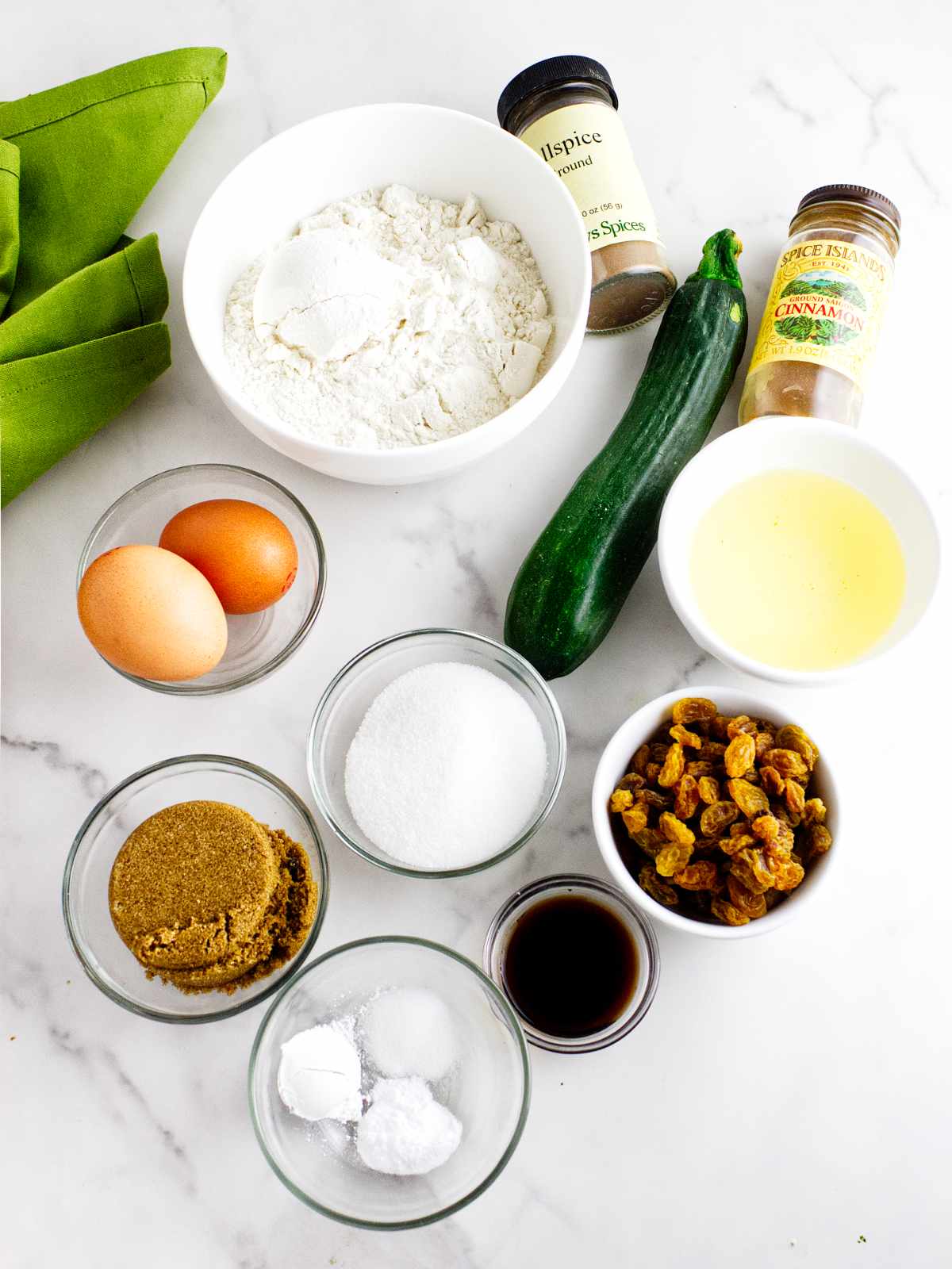 Various baking ingredients on a white surface, ready for making bread machine zucchini bread—flour, a whole zucchini, eggs, sugars, baking soda, vanilla, golden raisins, oil, allspice, and cinnamon. Green cloth in the corner adds a pop of color.