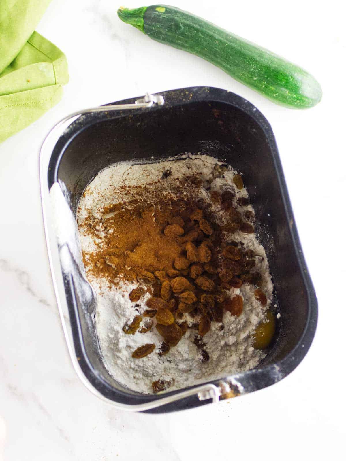 A bread machine pan filled with flour, raisins, cinnamon, and other ingredients for bread machine zucchini bread sits on a white surface next to a fresh zucchini and a green cloth.