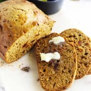 A loaf of bread machine zucchini bread with visible bits inside is partially sliced on a white surface. Two slices are in front, one with melting butter spread on top.