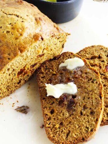 A loaf of bread machine zucchini bread with visible bits inside is partially sliced on a white surface. Two slices are in front, one with melting butter spread on top.