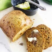 A loaf of bread machine zucchini bread sits on a cutting board with two slices cut, one spread with melting butter. Nearby are a small dish of butter with a knife and a fresh zucchini.