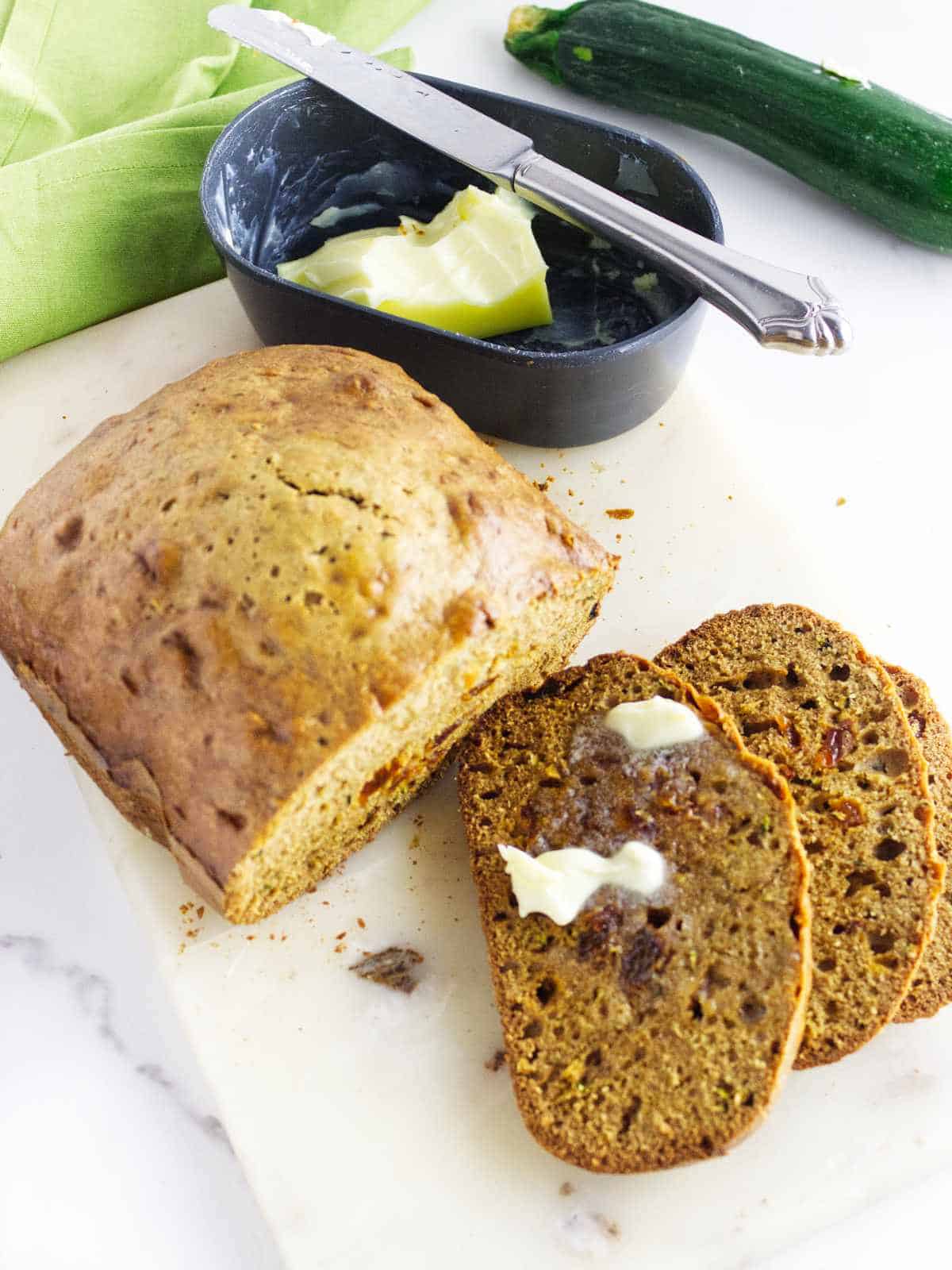 A loaf of bread machine zucchini bread sits on a cutting board with two slices cut, one spread with melting butter. Nearby are a small dish of butter with a knife and a fresh zucchini.