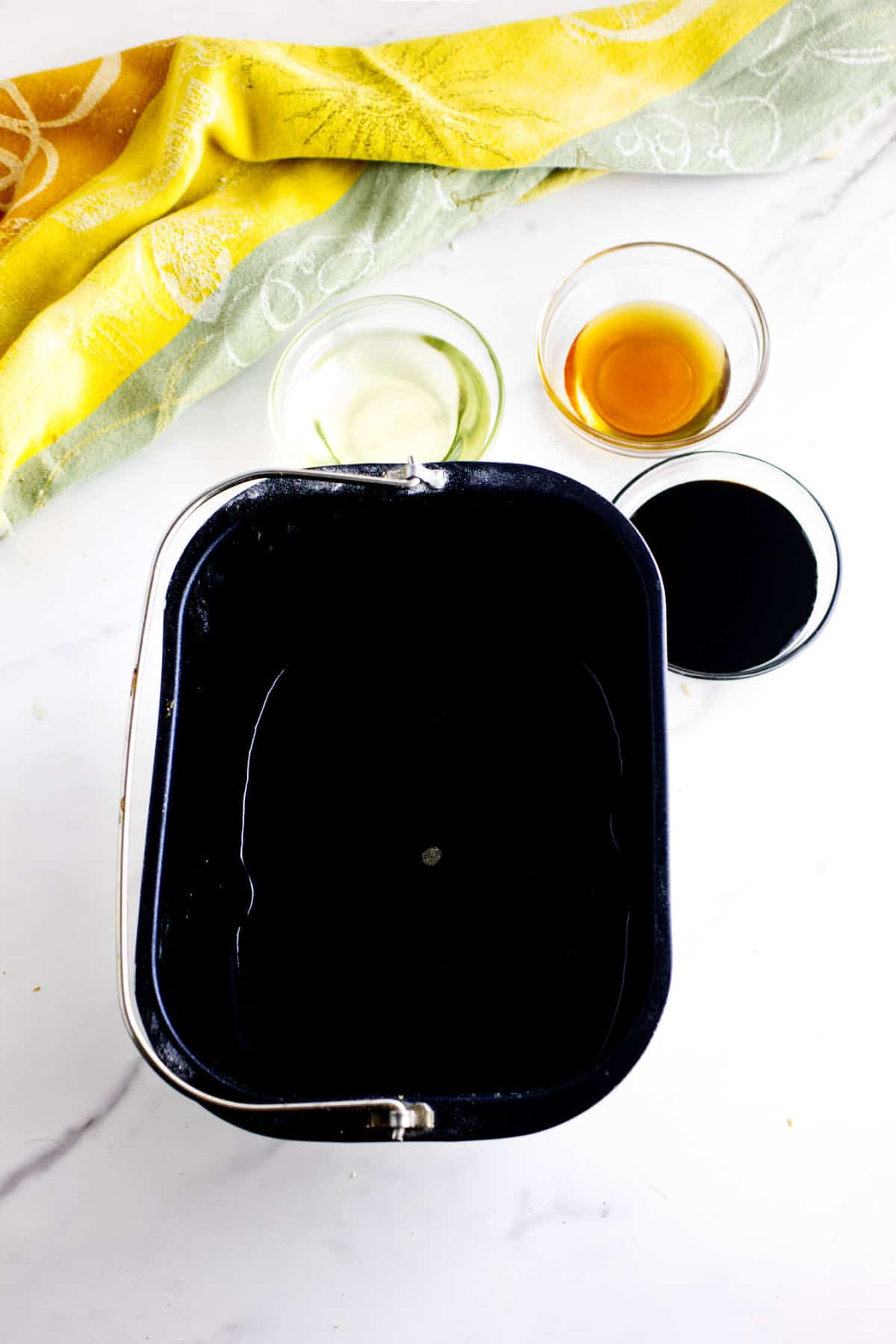 A black bread machine pan filled with water sits on a white surface next to small bowls of oil, vanilla extract, and another dark liquid-perfect for making Cheesecake Factory bread. A yellow and green patterned towel is in the background.