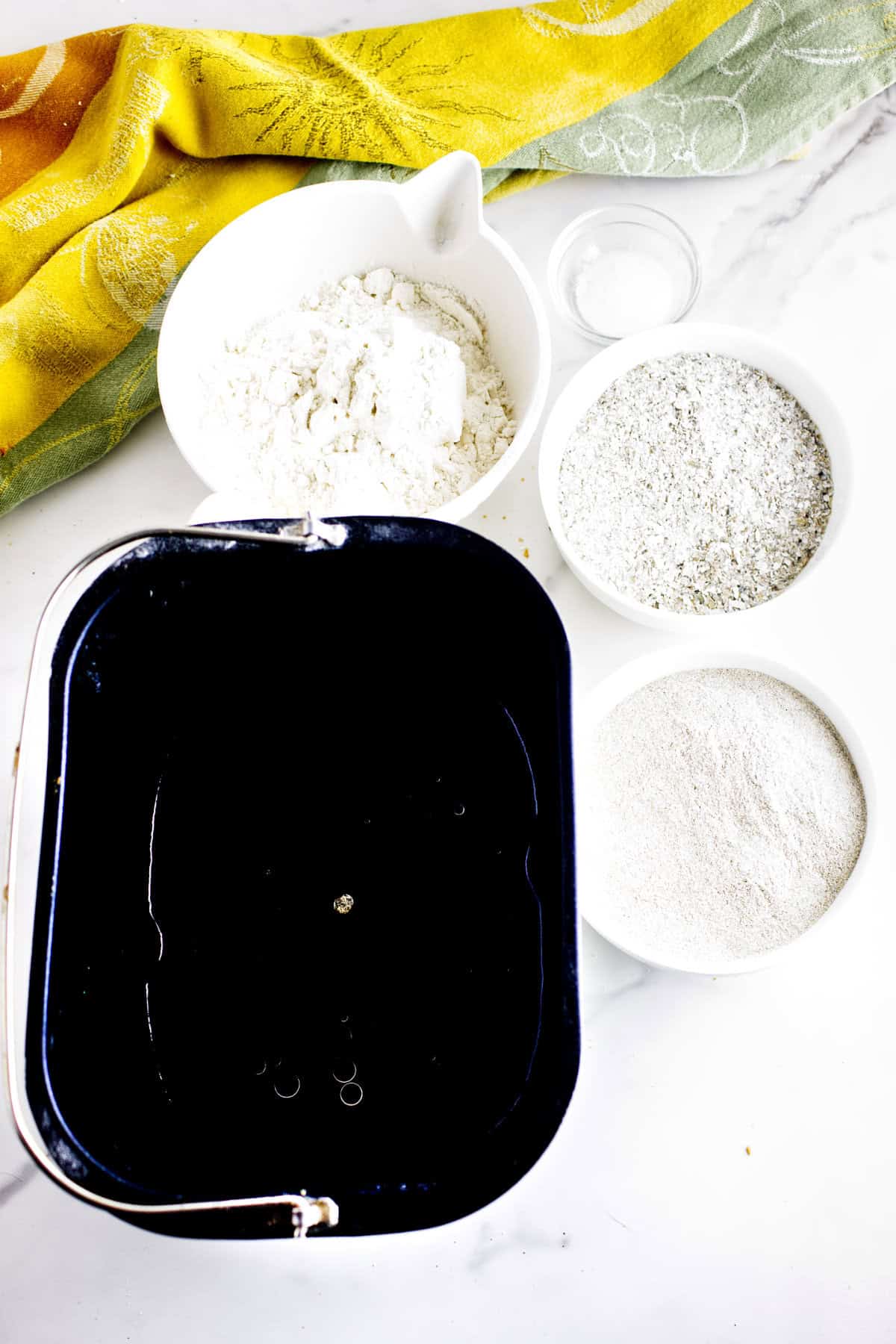 Four bowls on a white surface: one with flour, salt, oats, and another powdery ingredient-perfect for making bread machine Cheesecake Factory bread-alongside a black rectangular container and a yellow patterned cloth.