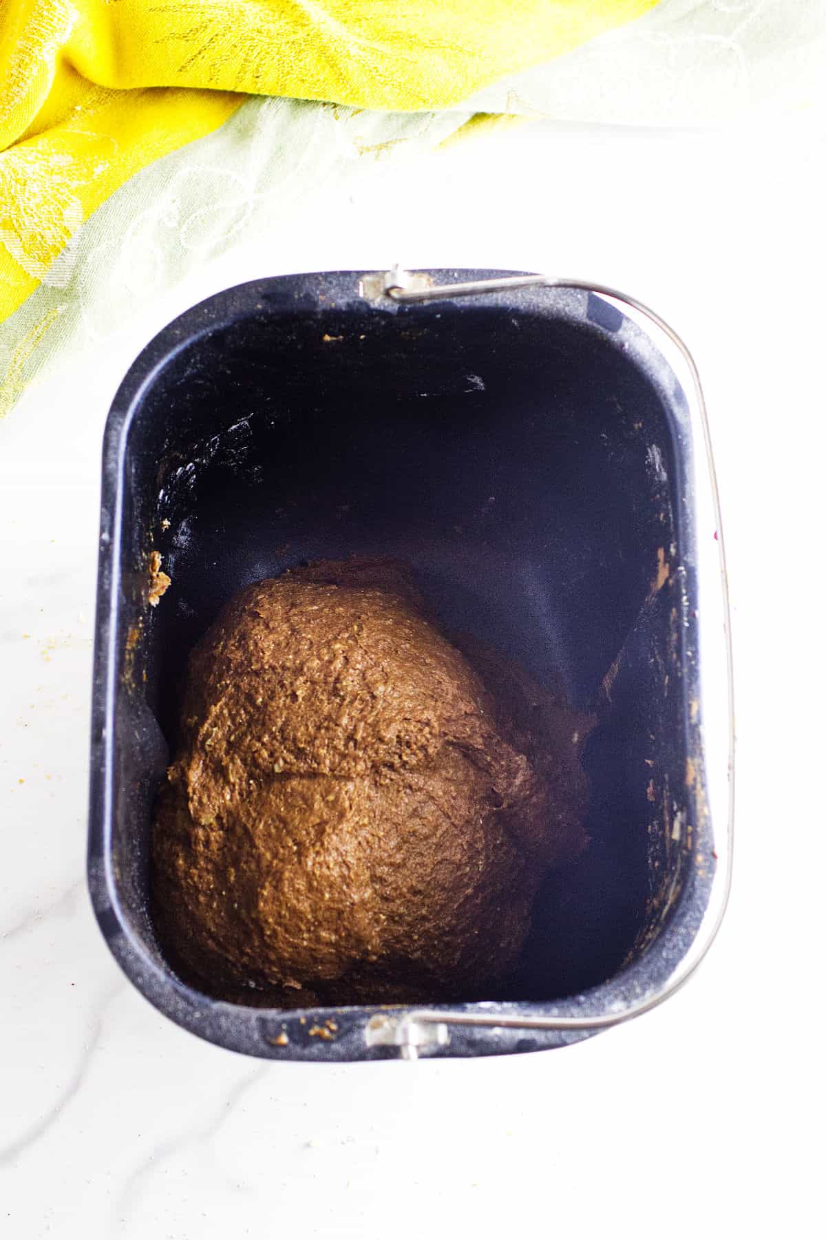A ball of brown bread machine cheesecake factory bread dough sits inside a black bread machine pan, with a yellow cloth partially visible in the background on a white countertop.