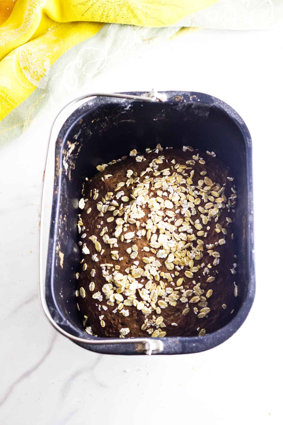 A black bread machine pan filled with Cheesecake Factory bread dough topped with oats, placed on a white surface near a yellow cloth.