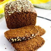 A loaf of dark brown bread, reminiscent of Cheesecake Factory bread, topped with oats and partially sliced on a white surface, with two slices in front. A yellow cloth is in the background.