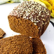A loaf of dark brown oat bread, partially sliced like cheesecake factory bread, with oat flakes on top. Two slices are lying in front of the loaf. A yellow and green cloth is in the background.