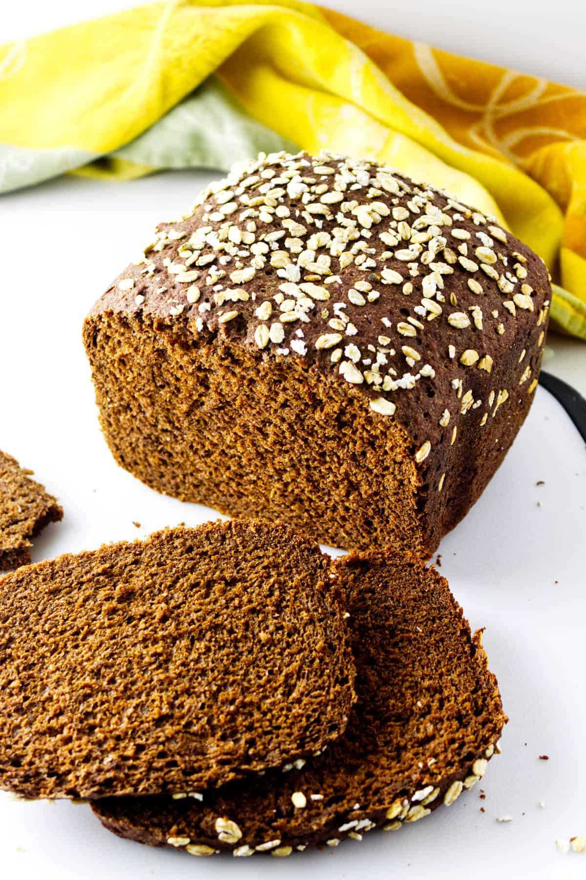A loaf of dark brown oat bread, partially sliced like cheesecake factory bread, with oat flakes on top. Two slices are lying in front of the loaf. A yellow and green cloth is in the background.