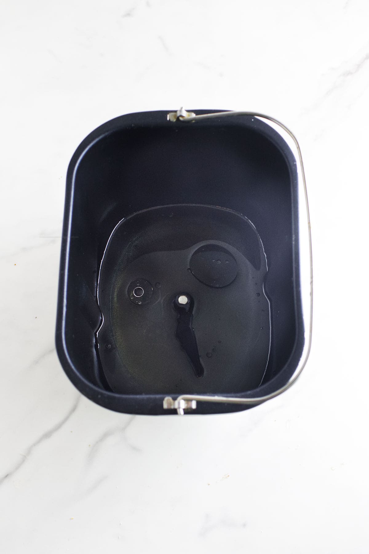 A black bread machine pan with a paddle in the center, partially filled with water, sits on a white marble surface-ready to help you create the perfect bread maker cinnamon swirl bread.