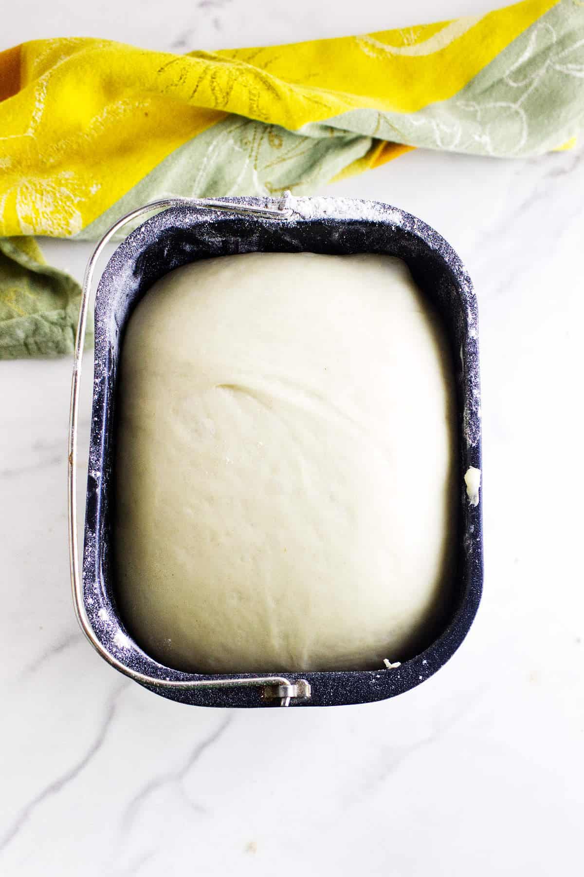 A metal bread machine pan filled with smooth, risen dough for cinnamon swirl bread sits on a white marble surface next to a yellow and green kitchen towel.