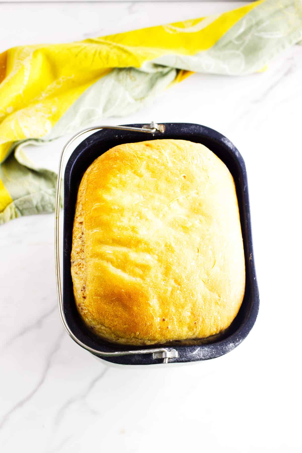 Golden brown loaf of bread maker cinnamon swirl bread in a bread machine pan on a white marble countertop, with a yellow and green kitchen towel in the background.