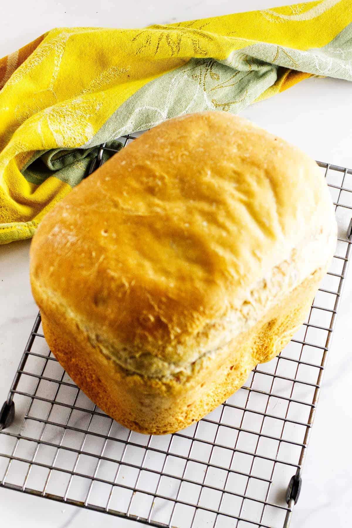 A golden-brown loaf of bread maker cinnamon swirl bread cools on a wire rack, with a yellow and green kitchen towel in the background on a white surface.