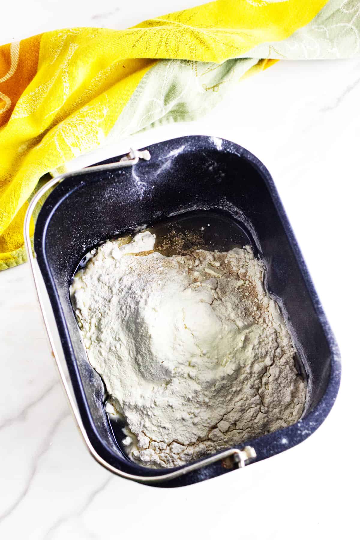 Bread machine pan filled with ingredients for bread maker cinnamon swirl bread, ready to mix. A yellow and green patterned kitchen towel is partially visible beside the pan on a white surface.