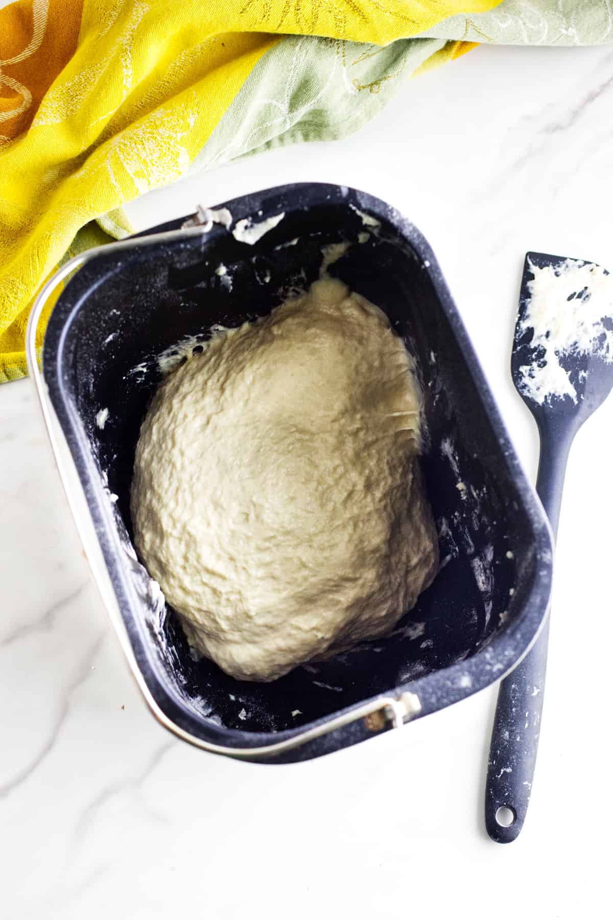 A bread machine pan filled with risen bread maker cinnamon swirl bread dough sits on a white surface next to a black spatula with dough residue; a yellow and green cloth is partially visible in the background.