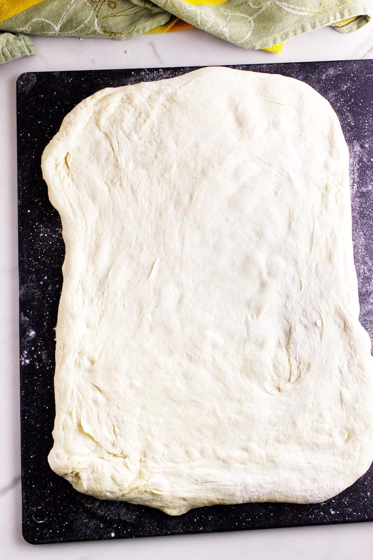 A large, rectangular piece of pizza dough-resembling bread maker cinnamon swirl bread-is stretched out on a dark, floured surface, ready for toppings. A patterned green cloth is partially visible at the top of the image.