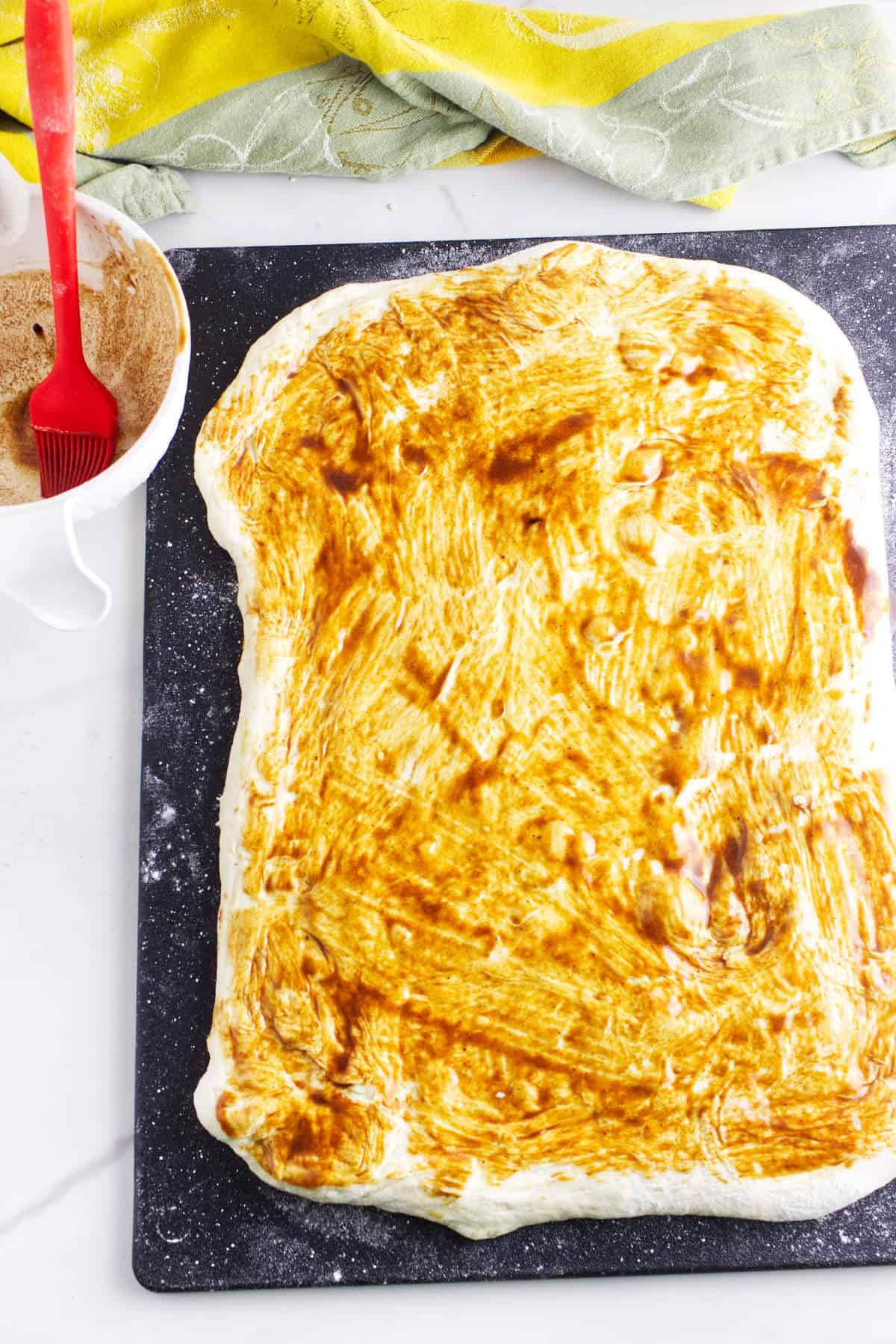 Rectangular rolled out dough on a black board coated with brown cinnamon mixture for bread maker cinnamon swirl bread, a red brush and white bowl of extra mixture nearby; green and yellow towels in the background.