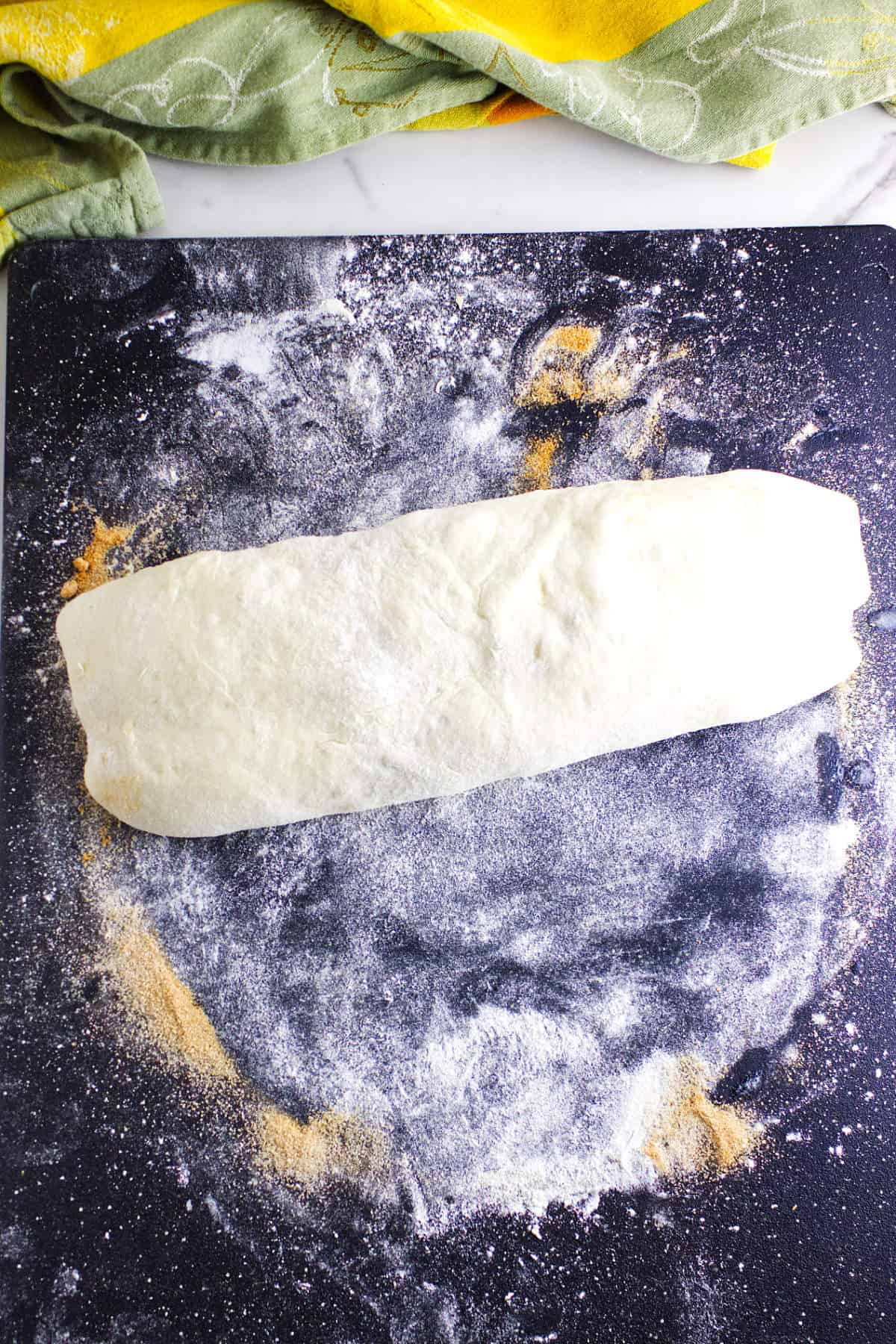 A rolled log of dough, destined to become bread maker cinnamon swirl bread, sits on a floured black surface with scattered flour and cornmeal; a green and yellow cloth is partially visible at the top of the image.