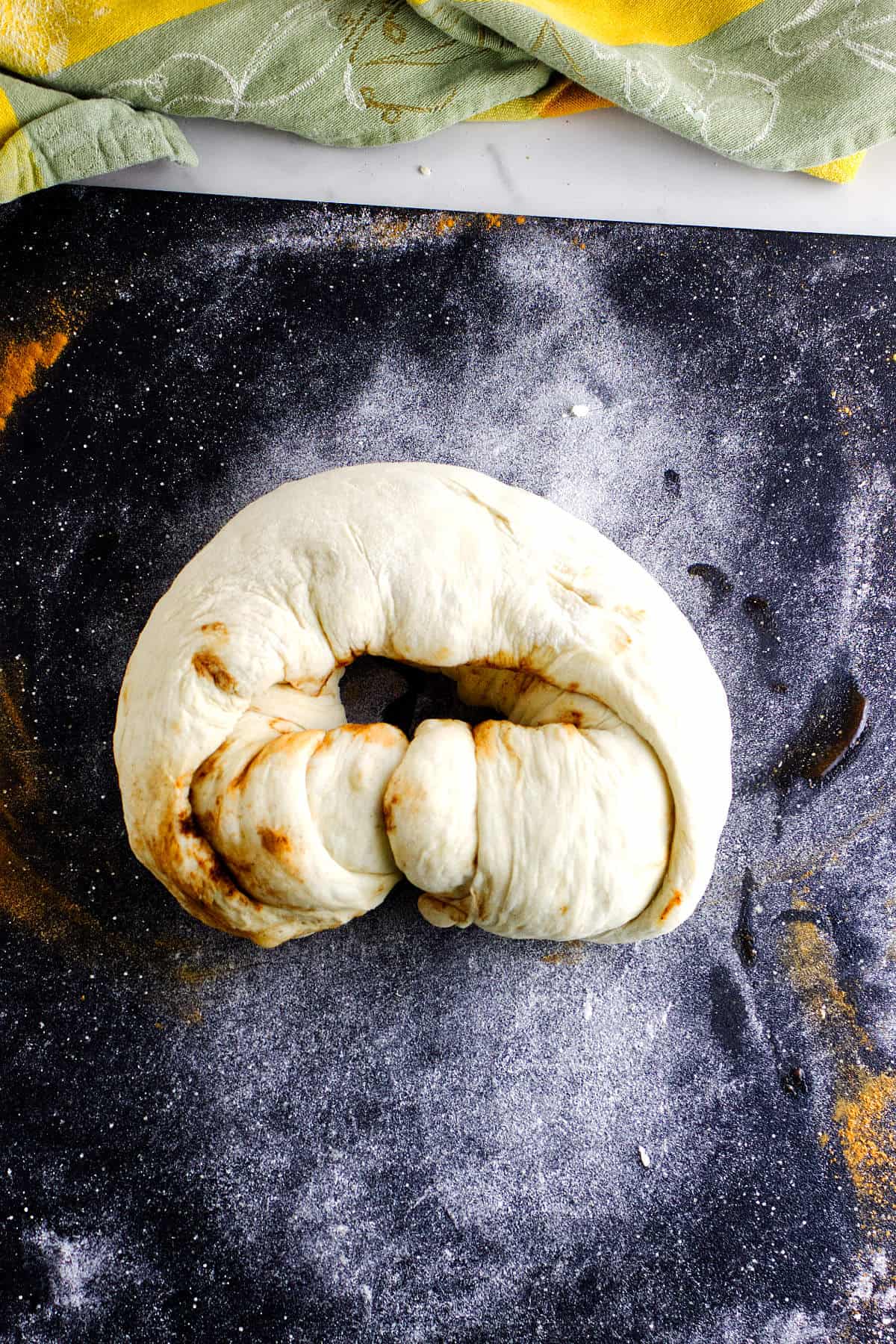 A piece of unbaked, twisted dough for bread maker cinnamon swirl bread sits on a floured, dark surface with a yellow and green cloth partially visible in the background.