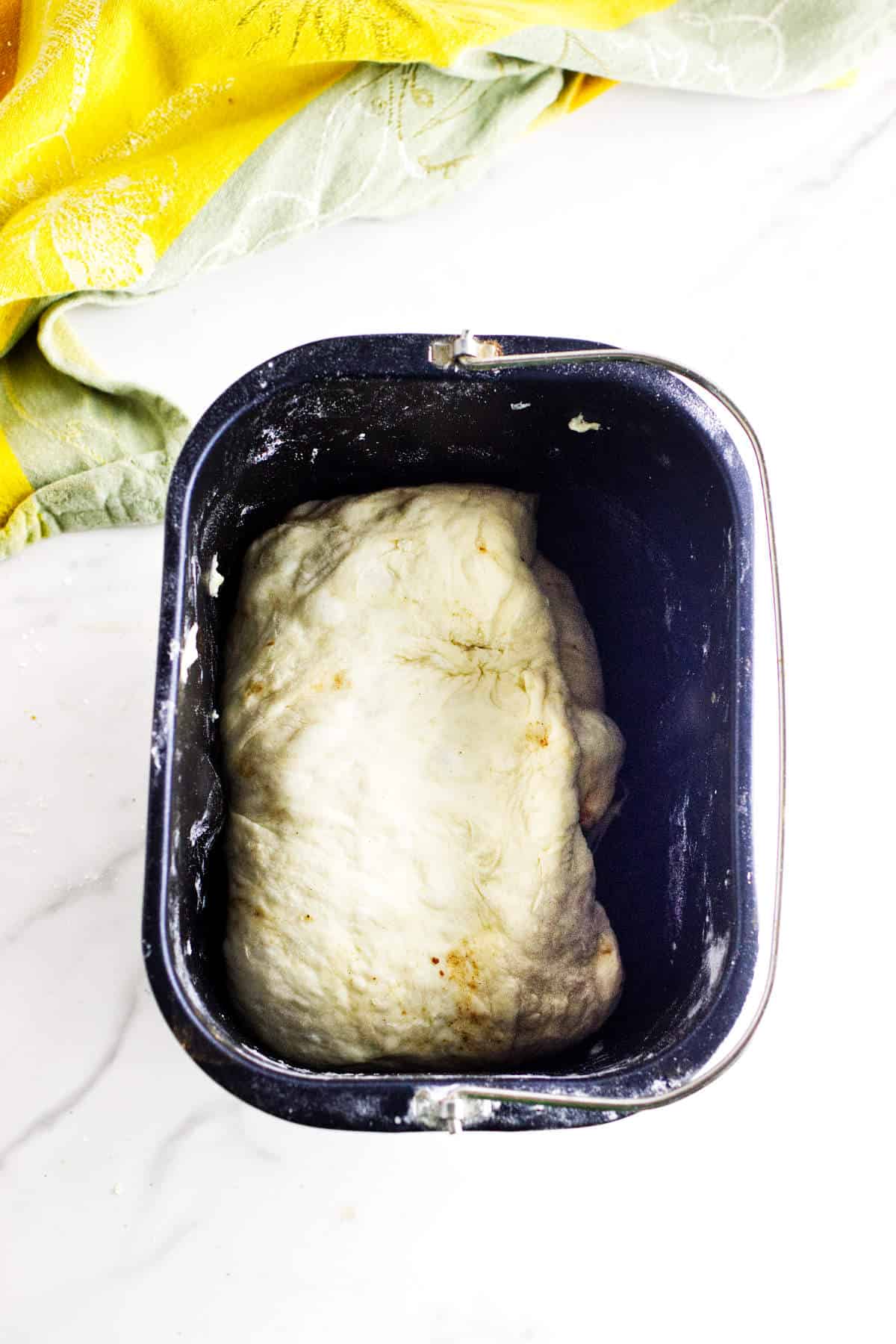 A ball of dough for bread maker cinnamon swirl bread rests inside the bread machine pan, with part of a yellow and green cloth visible in the background on a white surface.