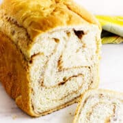 A close-up of bread maker cinnamon swirl bread with one slice cut, showing the spiral of cinnamon inside. The loaf rests on a white surface with a yellow and green cloth in the background.