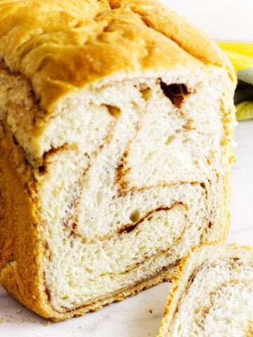 A close-up of bread maker cinnamon swirl bread with one slice cut, showing the spiral of cinnamon inside. The loaf rests on a white surface with a yellow and green cloth in the background.