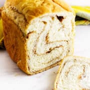 A loaf of bread maker cinnamon swirl bread with one slice cut off, showing the spiral pattern inside. The bread is on a white surface with a yellow cloth in the background.