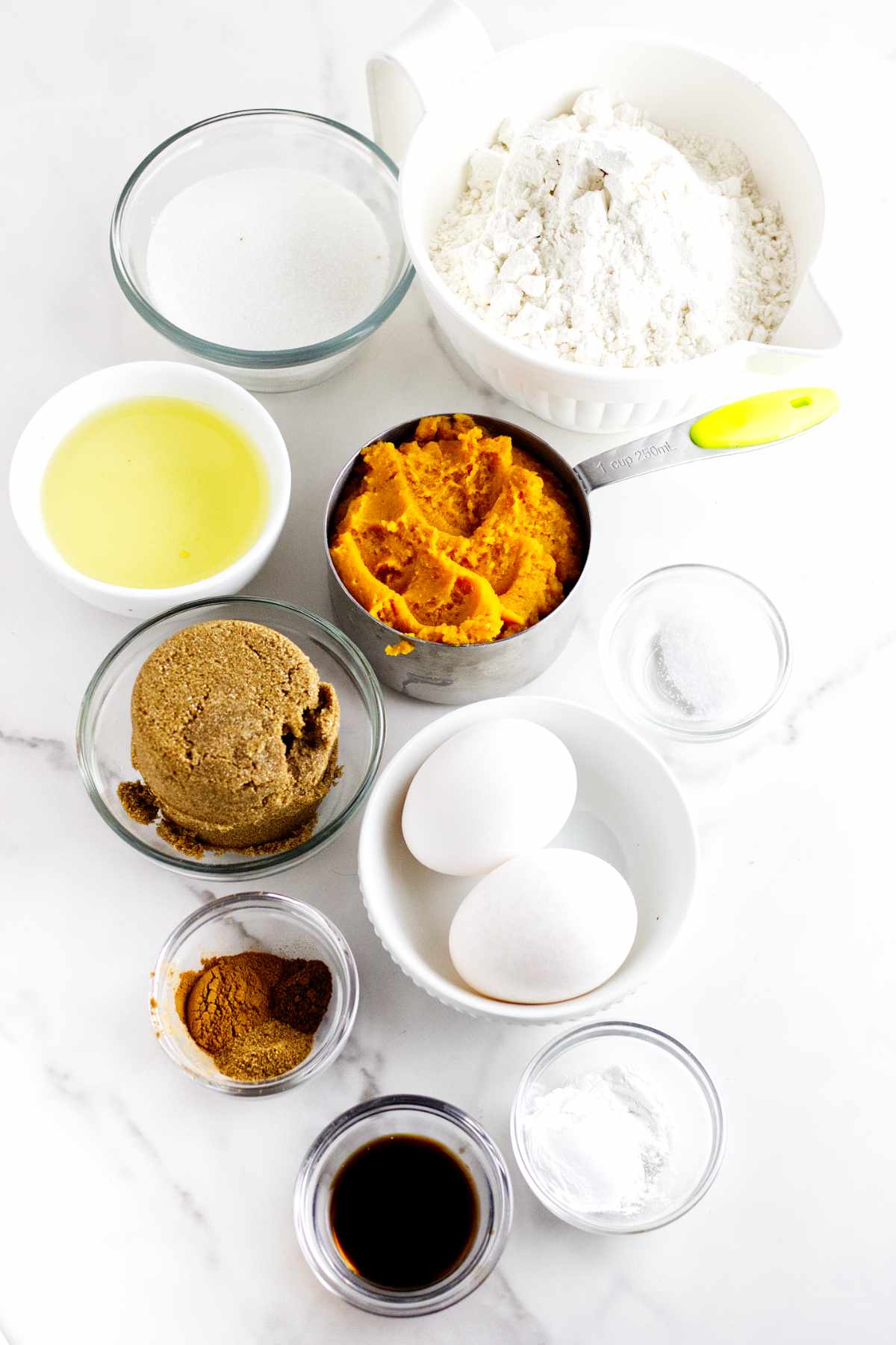 Top-down view of baking ingredients for bread machine pumpkin bread: flour, sugars, pumpkin puree, eggs, oil, vanilla extract, baking powder, baking soda, salt, and pumpkin pie spice on a white surface.
